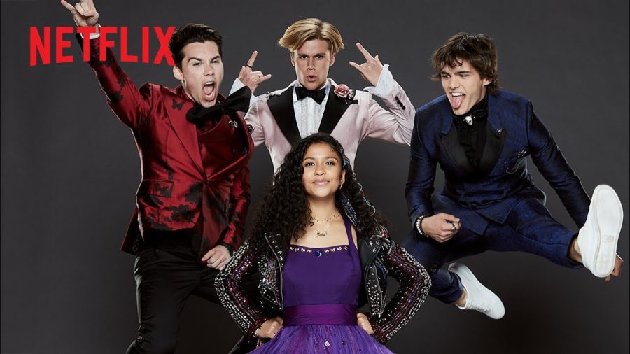 Boy in red suit jumps with his right arm in the air. Second boy in a light pink suit jumps up making the rock and roll symbol with both hands. Third boy in dark blue suit jumps up playing air guitar, kicking his left leg out. A girl in a purple dress stands below them, hands on her hips.